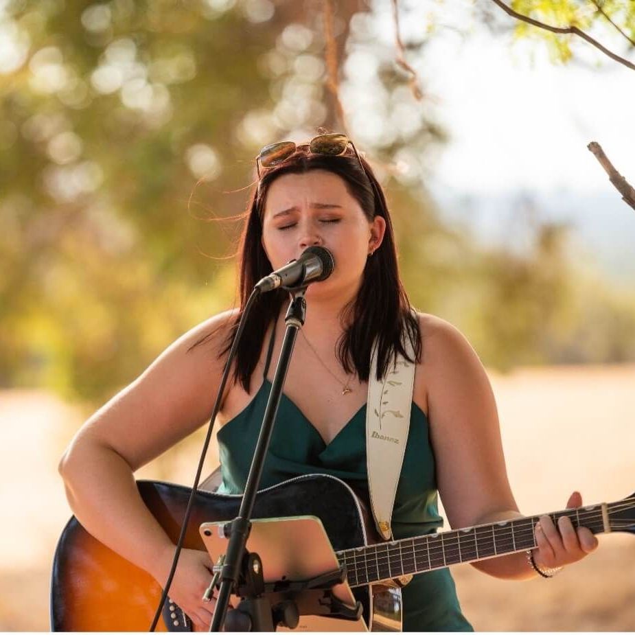 Woman singing and playing acoustic guitar outdoors, wearing a green dress.