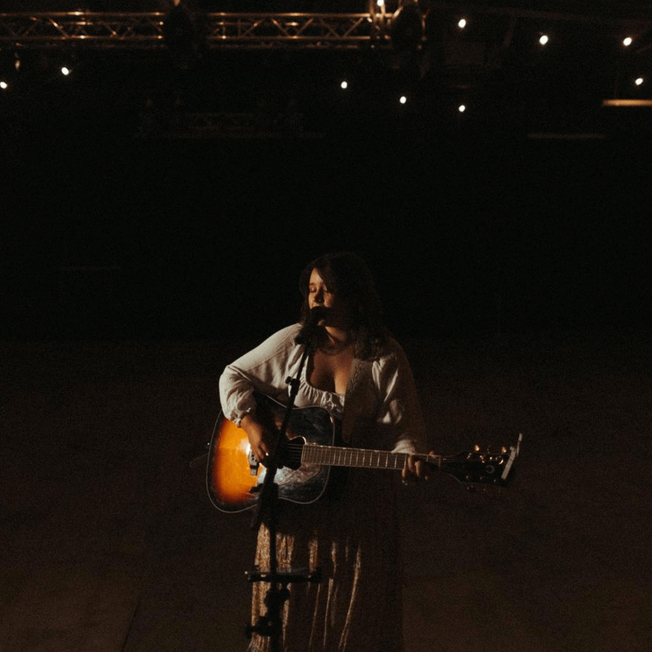 Woman playing an acoustic guitar on stage, lit by spotlights. Dark background.