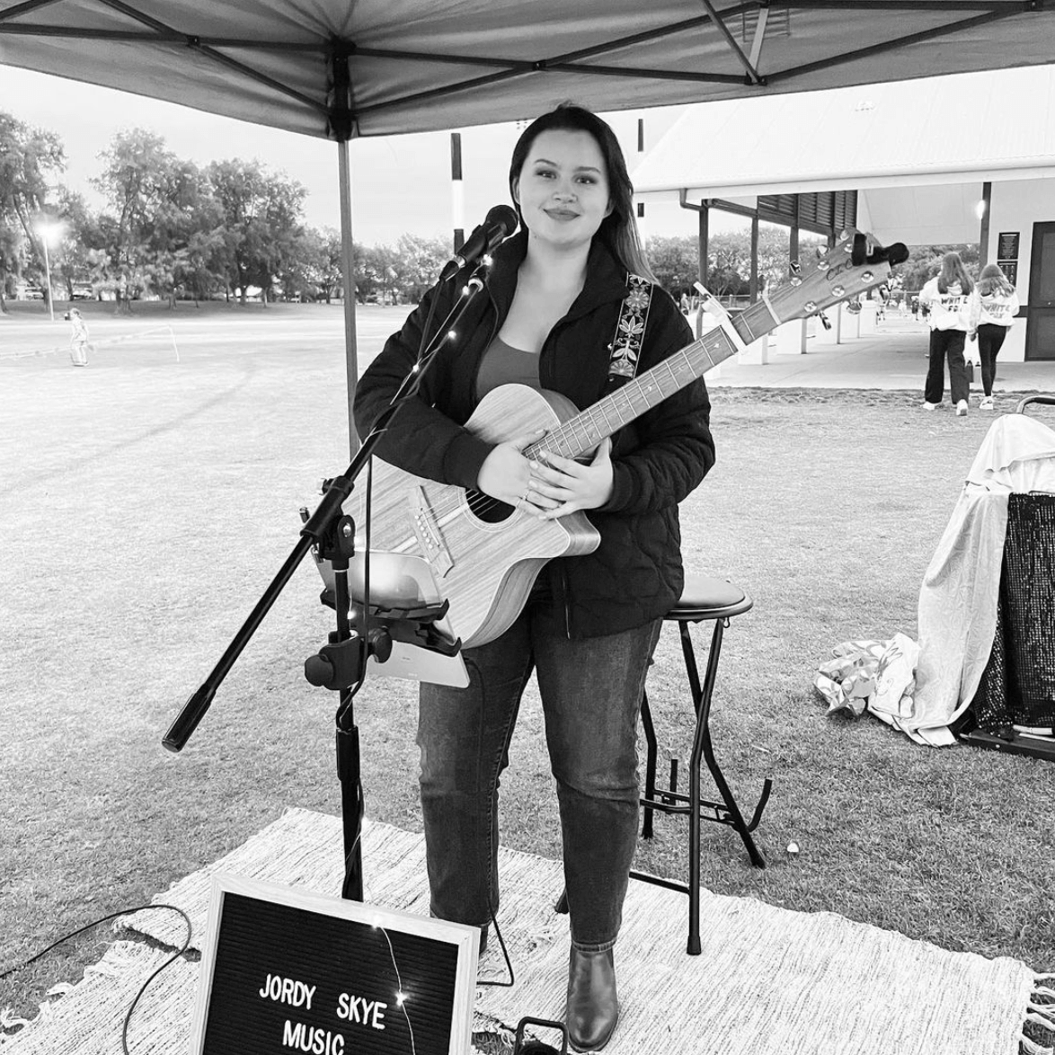 Woman playing an acoustic guitar and singing at an outdoor event, microphone in front of her.
