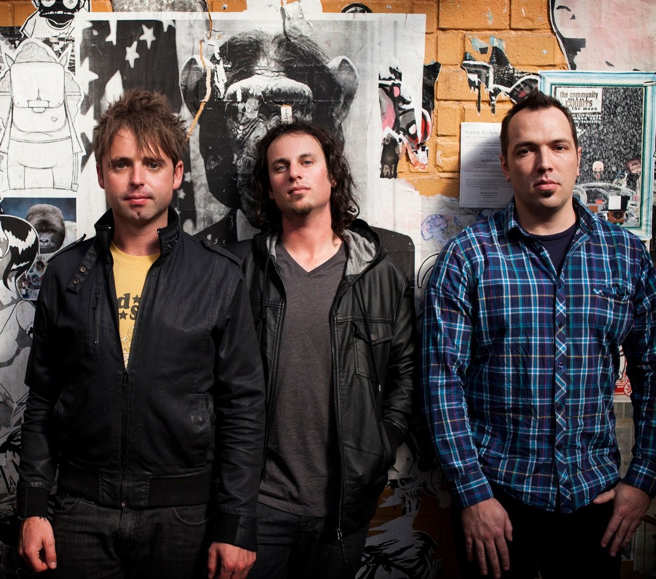 Three men stand side-by-side in front of a wall covered with black-and-white posters and artwork.
