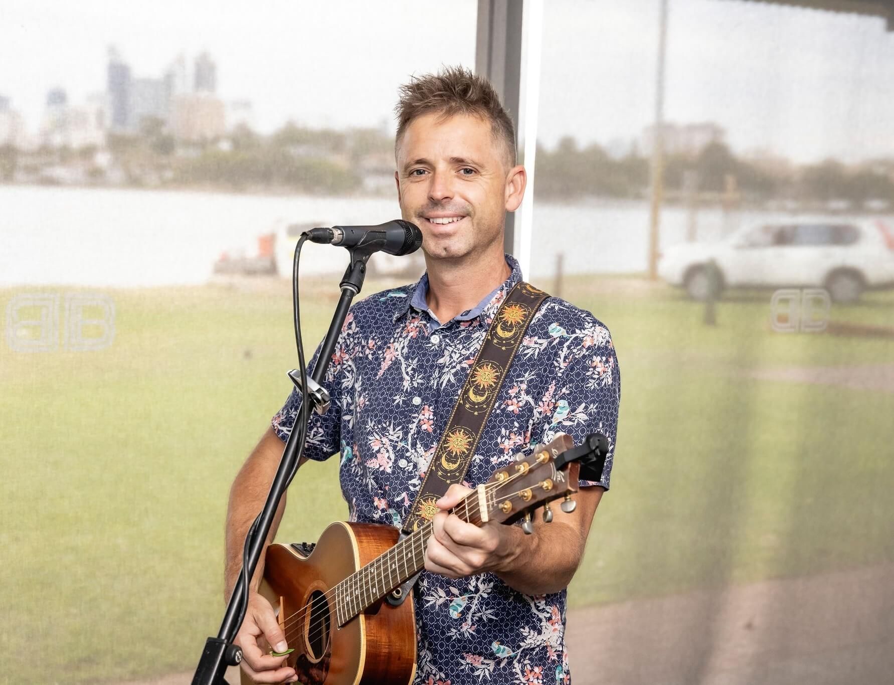 A person smiles while playing an acoustic guitar and singing into a microphone outdoors with a city skyline in the distance.