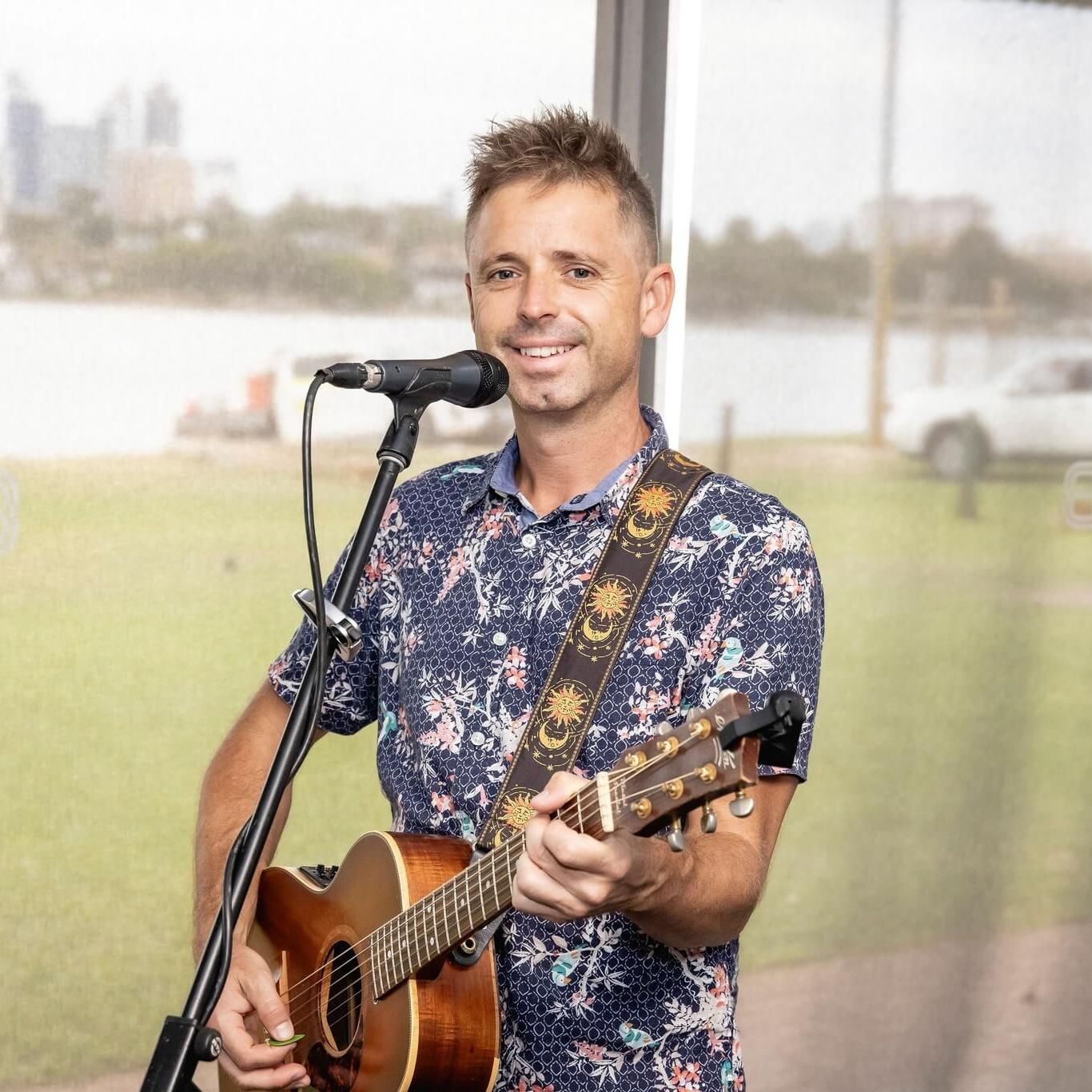 A musician with a patterned shirt playing an acoustic guitar and singing into a microphone outdoors by a waterfront.