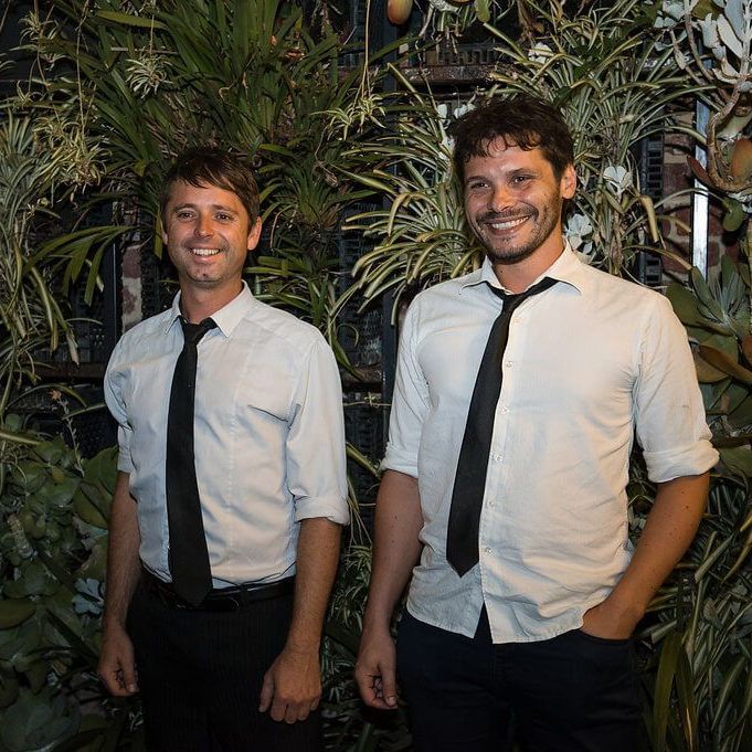 Two individuals wearing white button-up shirts and black ties stand side-by-side in front of a wall of green foliage.