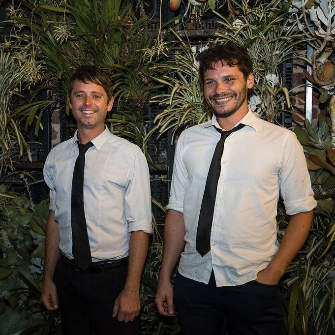 Two people in white button-down shirts and black ties stand side-by-side smiling in front of a dense wall of greenery.