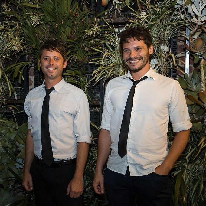 Two people wearing light-colored button-up shirts and dark ties, smiling in front of a lush, vertical wall of greenery.