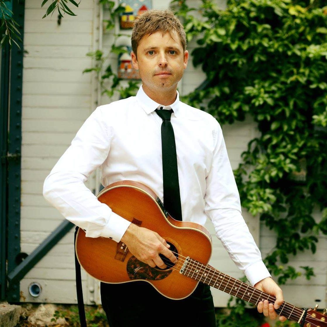 A person in a white button-down shirt and black tie holds an acoustic guitar while standing in front of a white wall.