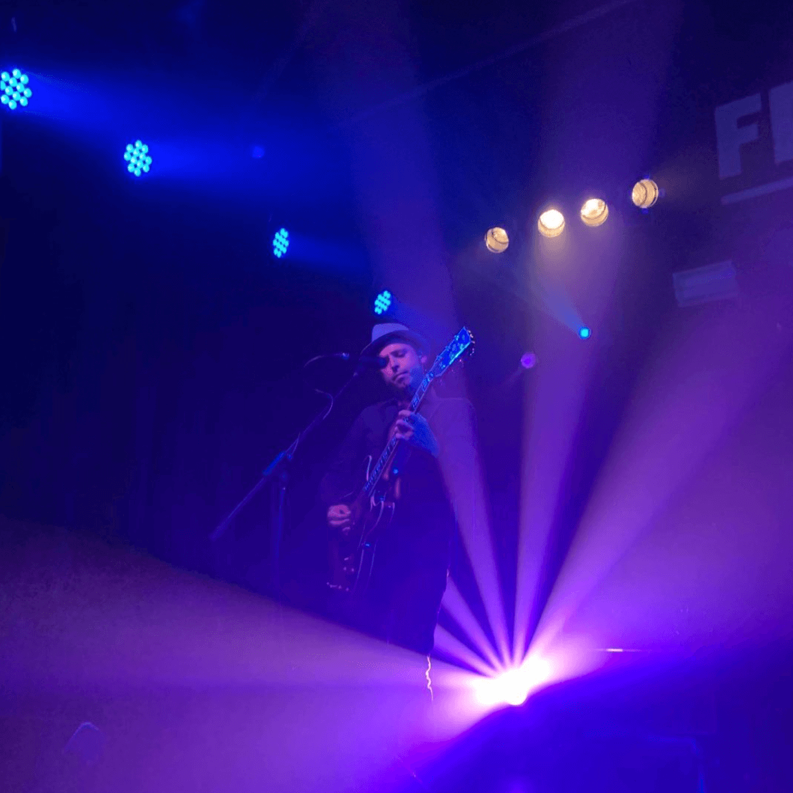 A musician plays an electric guitar on a dark stage illuminated by bright blue and purple spotlights.