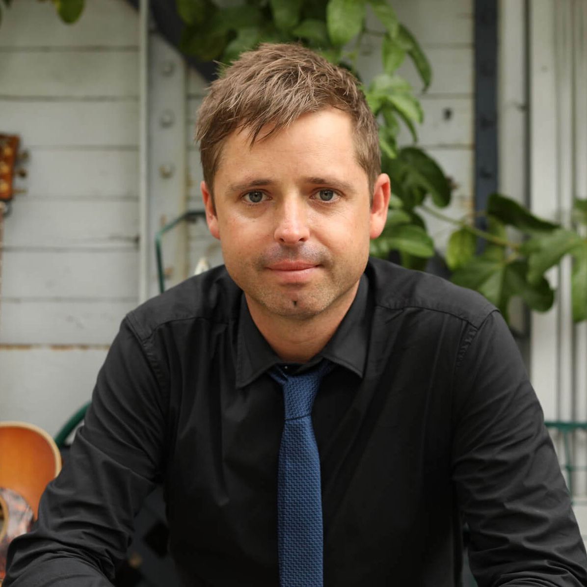 A person with short brown hair wears a black button-down shirt and blue tie, positioned before a white paneled background.
