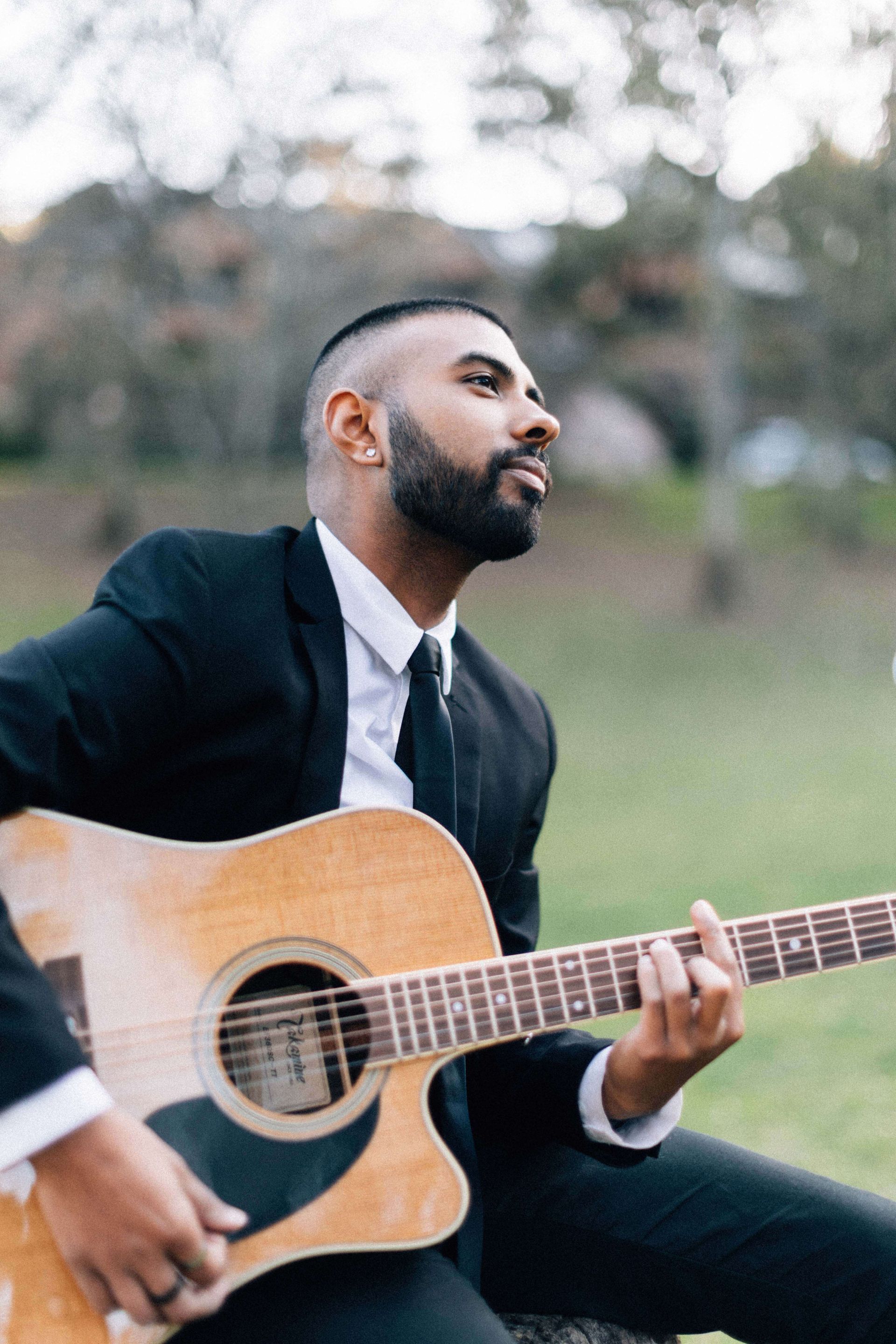 A person in a black suit and white shirt plays an acoustic guitar outdoors in a field with trees in the background.