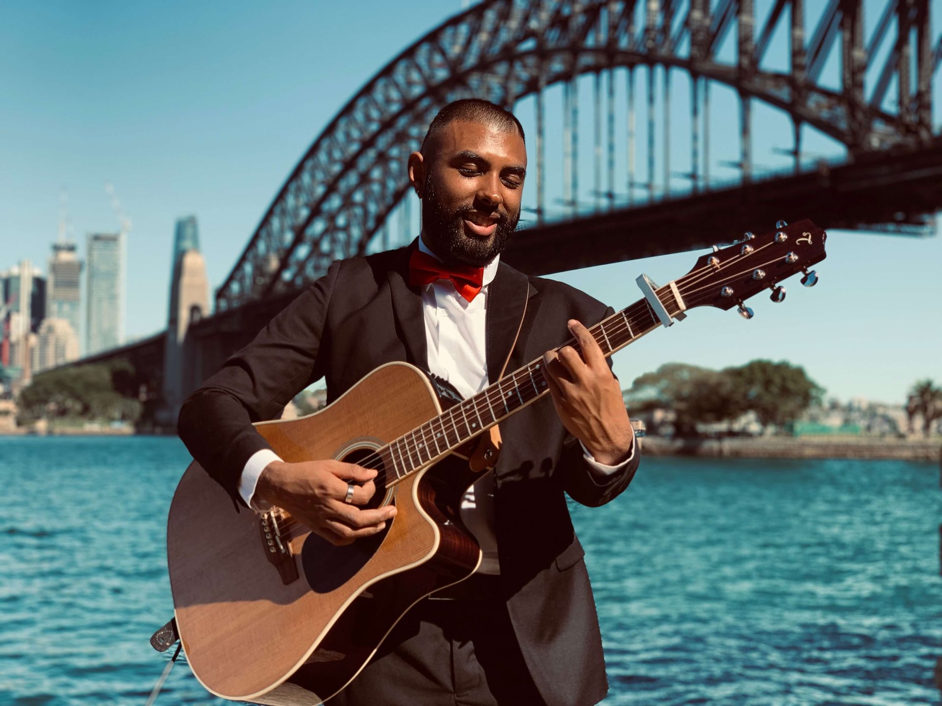 A person in a suit and red bow tie plays an acoustic guitar outdoors by the water with a large bridge in the background.
