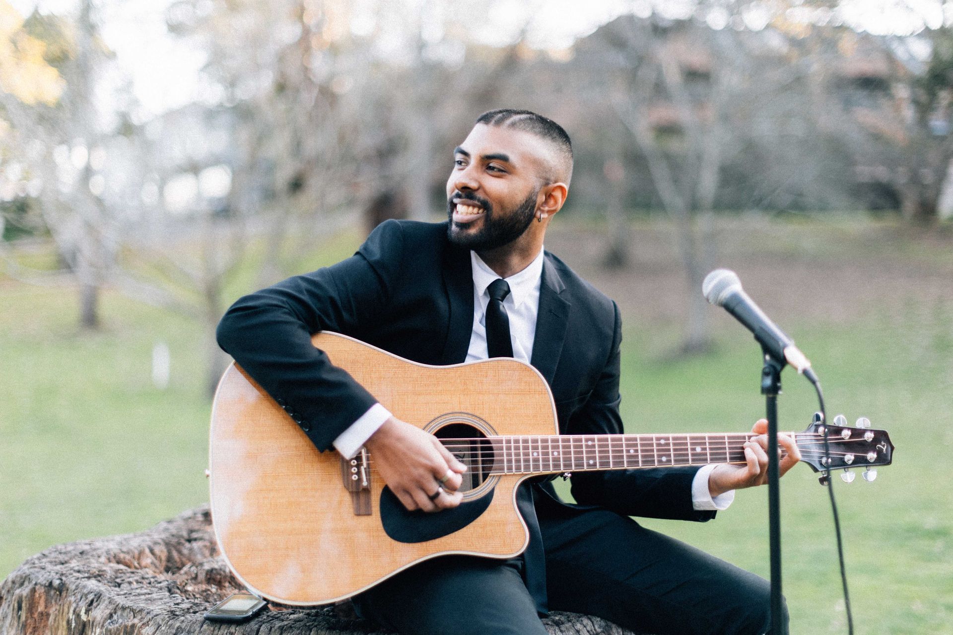 A person in a suit sits on a stump outdoors, playing an acoustic guitar and singing into a nearby microphone.