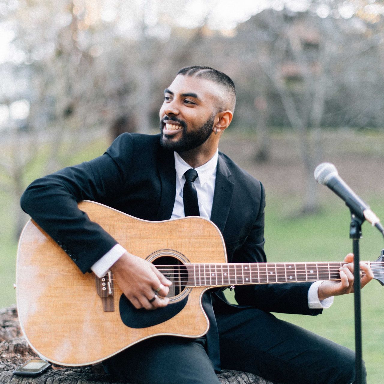 A person in a black suit and tie smiling while playing an acoustic guitar next to a microphone in an outdoor setting.