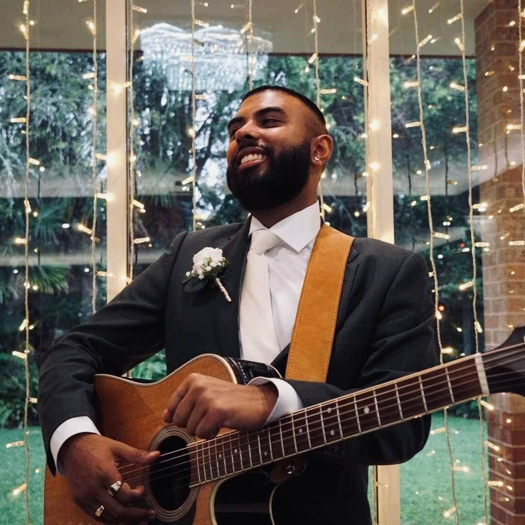 A person in a suit and tie playing an acoustic guitar in front of a window decorated with warm string lights.