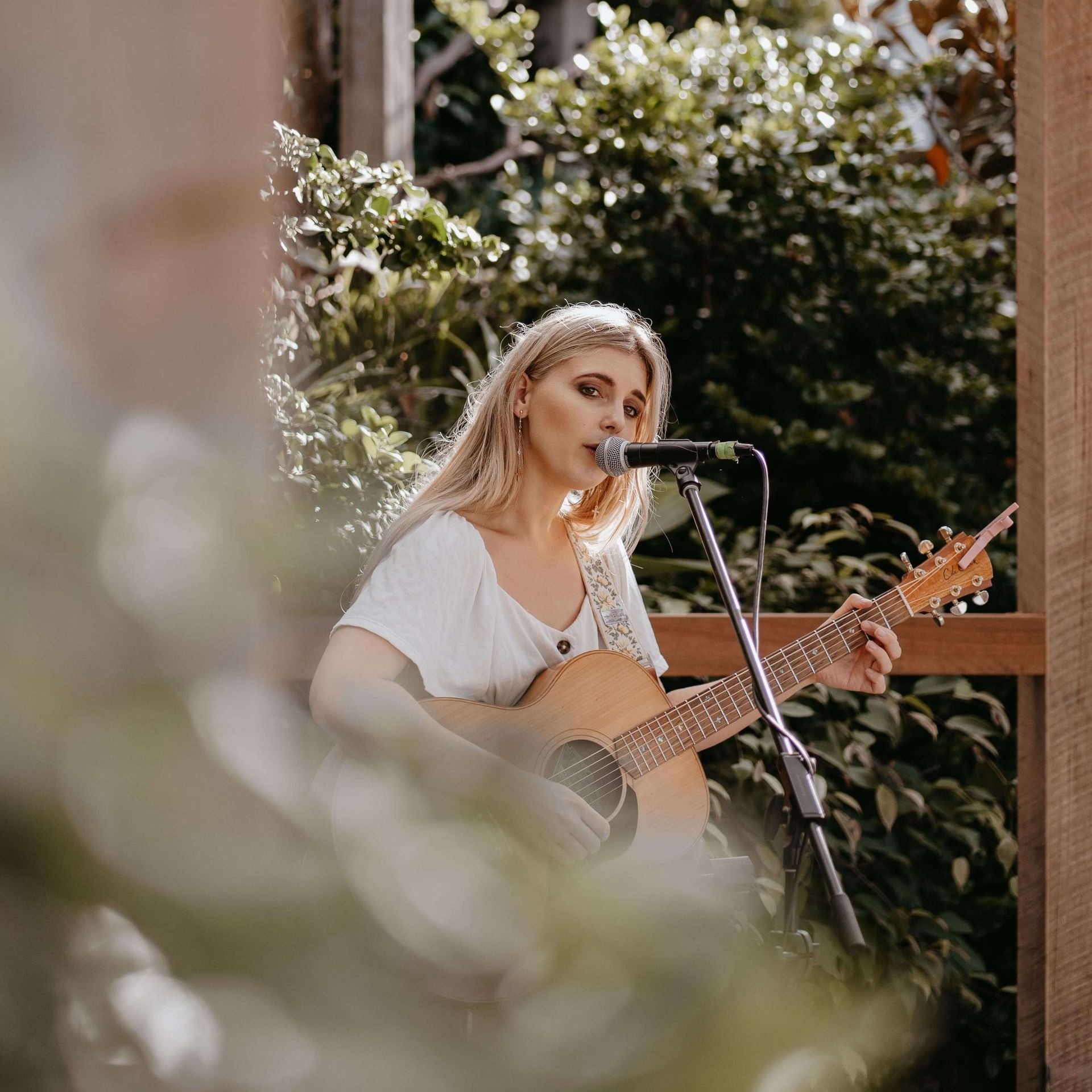 A person with long hair playing an acoustic guitar and singing into a microphone outdoors among lush green foliage.