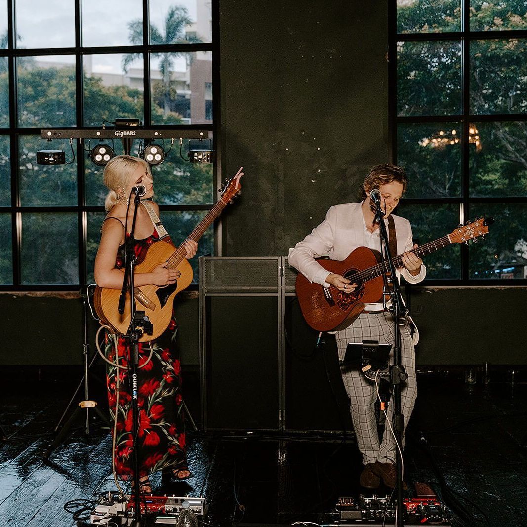 Two musicians stand side-by-side on a stage performing with acoustic guitars, positioned in front of large windows.