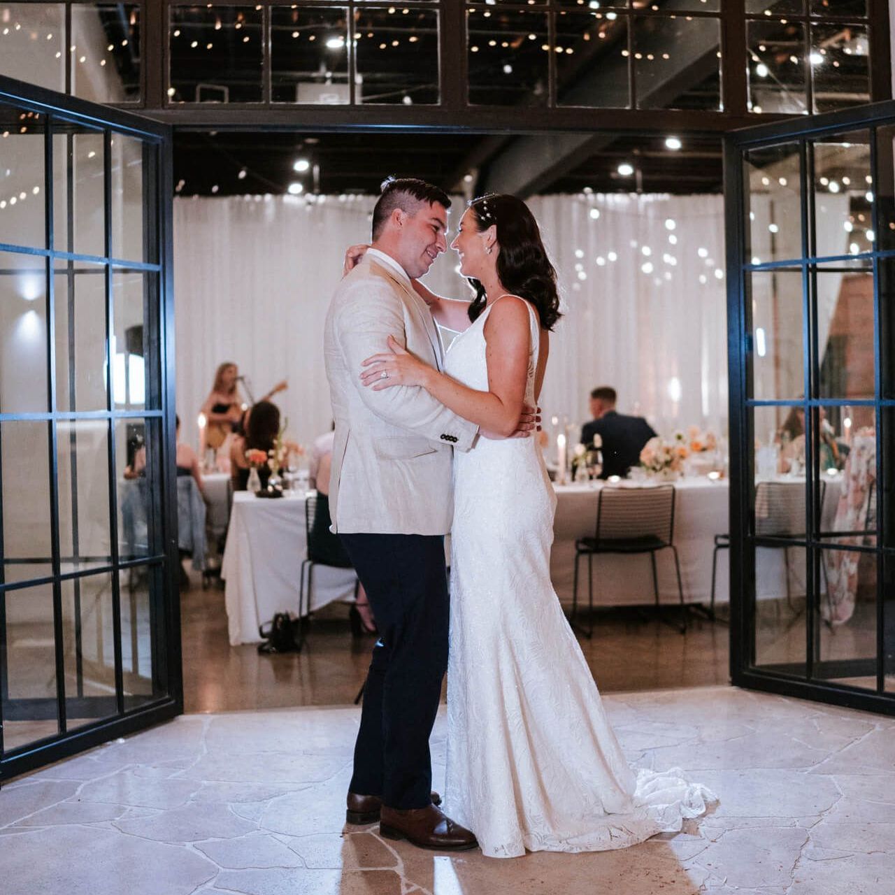 A couple dances during a wedding reception, framed by large glass doors with an softly lit dining area in the background.