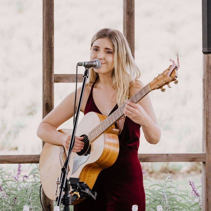 Woman singing into a microphone while playing acoustic guitar on an outdoor stage