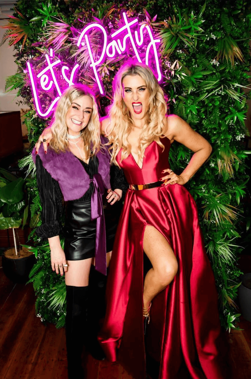 Two women pose in front of a neon 
