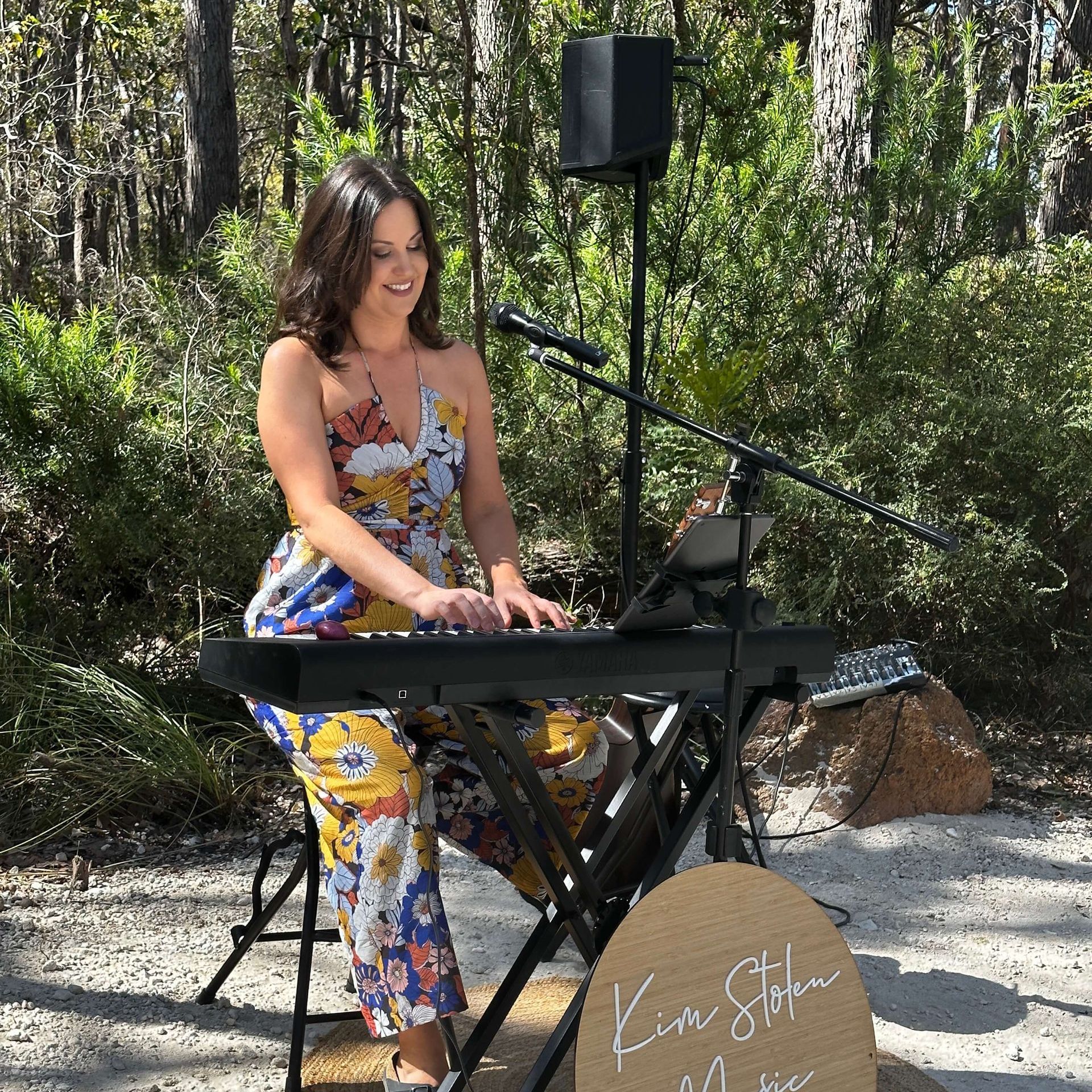 Woman playing a keyboard outside; microphone, speaker, and small sign in view. She wears a floral jumpsuit and smiles.