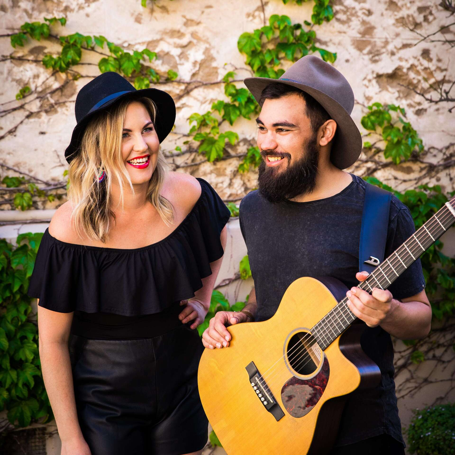 A smiling duo in black outfits stands before a vine-covered wall. Man holds a guitar.