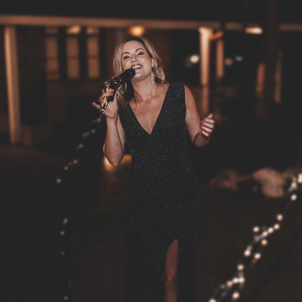 Woman in a black dress singing into a microphone outdoors at night, smiling, with string lights.