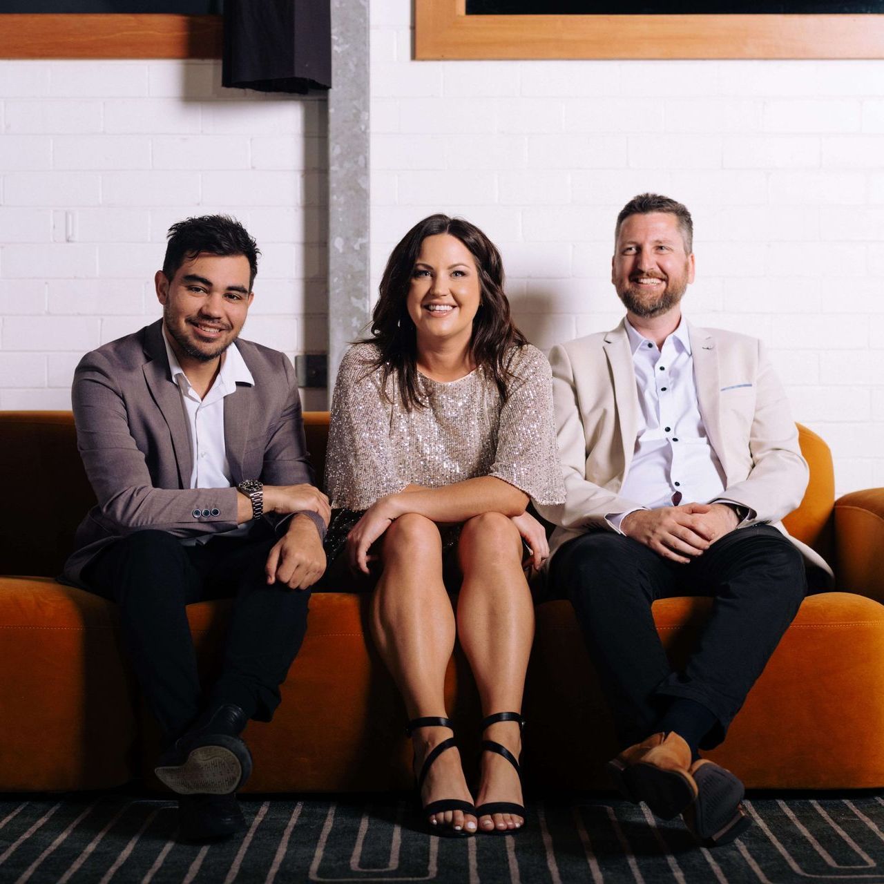 Three people sit on an orange couch. Woman in sparkly dress flanked by men in jackets. Modern setting.