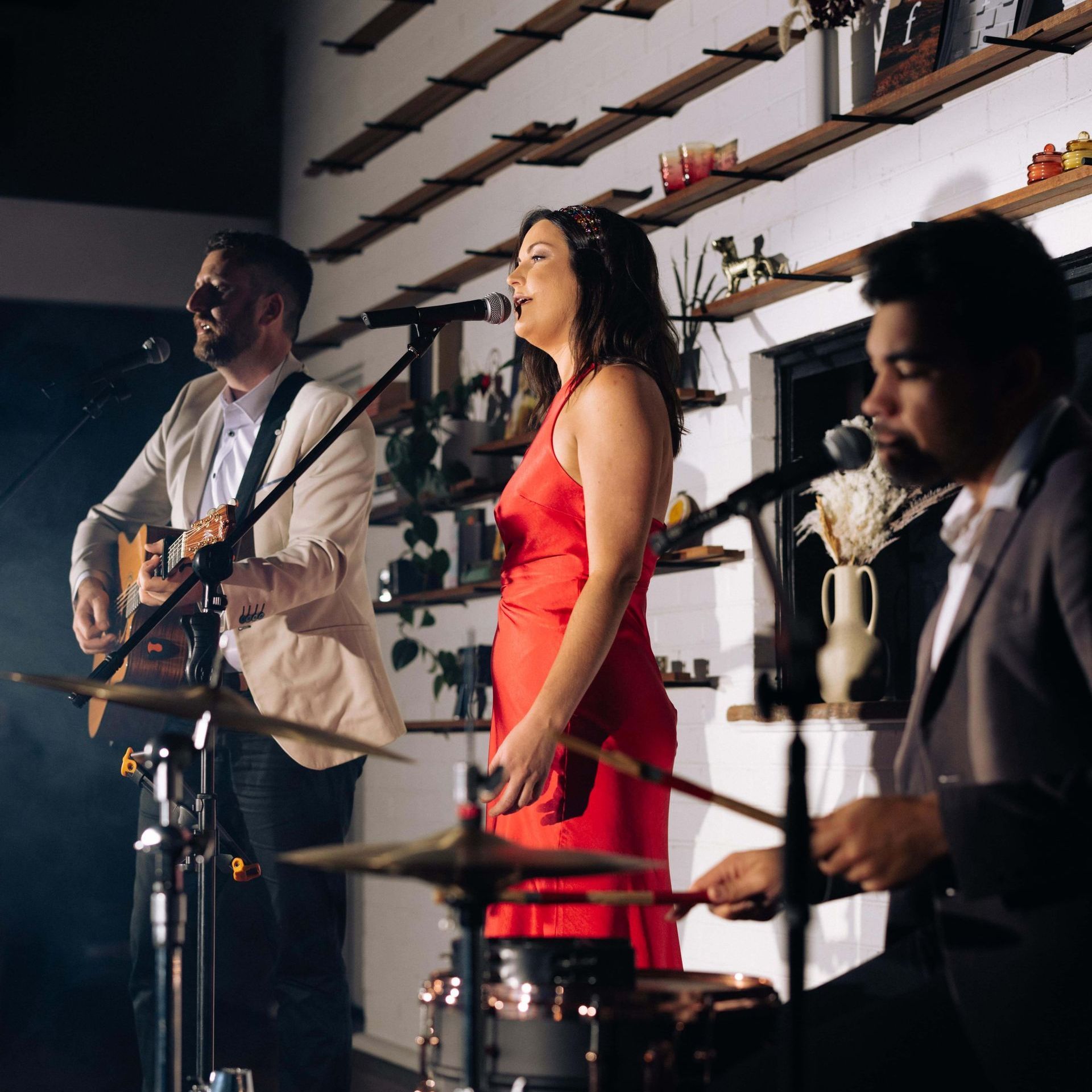 Band performing live on stage: guitarist, vocalist in red dress, drummer. Stage lit with warm lights.