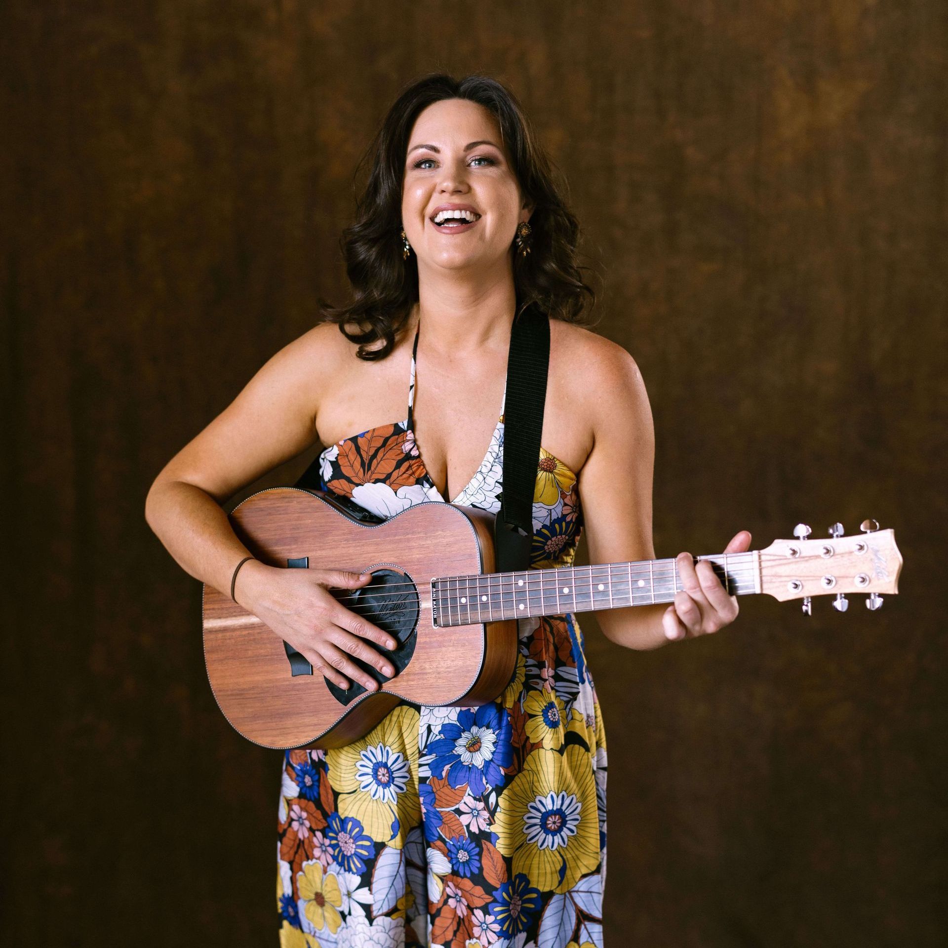 Woman smiling, playing acoustic guitar, wearing floral jumpsuit. Brown background.