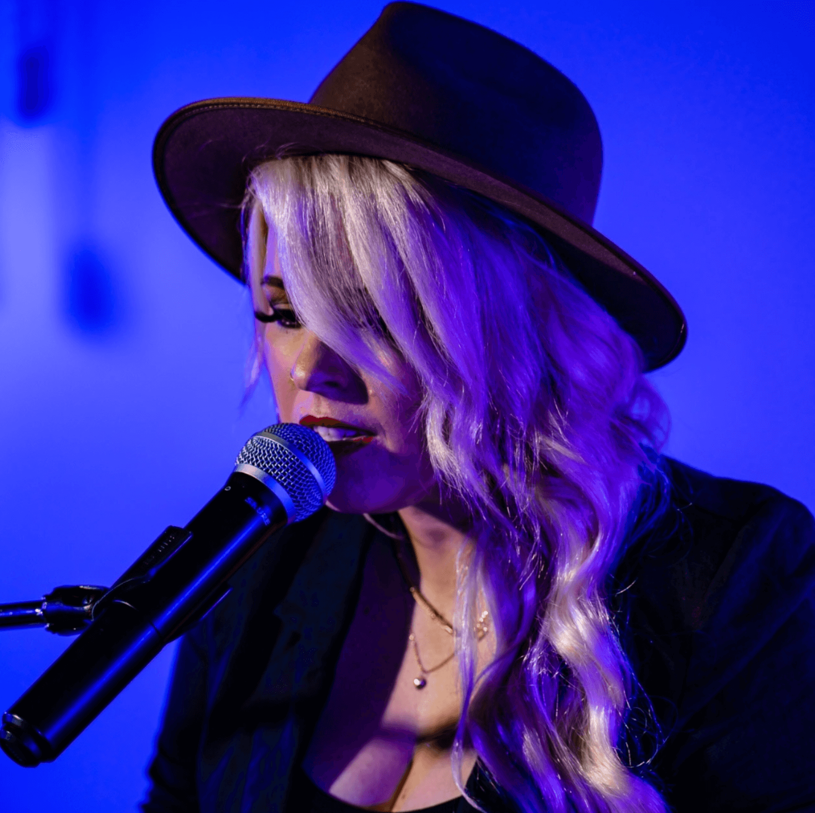 Woman with blonde hair wearing a fedora, singing into a microphone. Blue background.