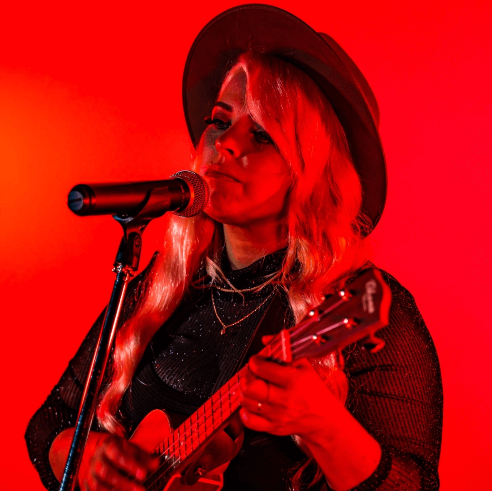 Woman in hat playing ukulele, singing into microphone under red stage lighting.