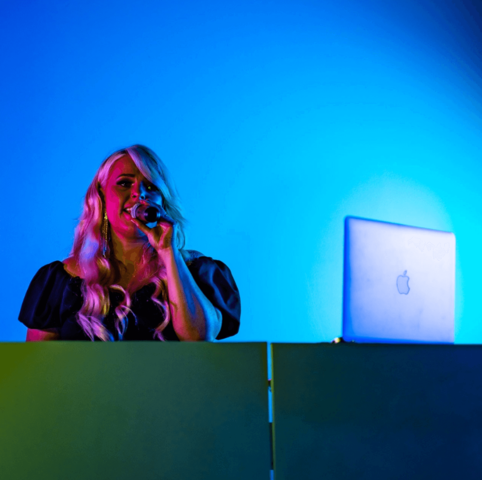 Woman with blonde hair singing into a microphone, standing behind a DJ booth with a laptop against a blue lit background.