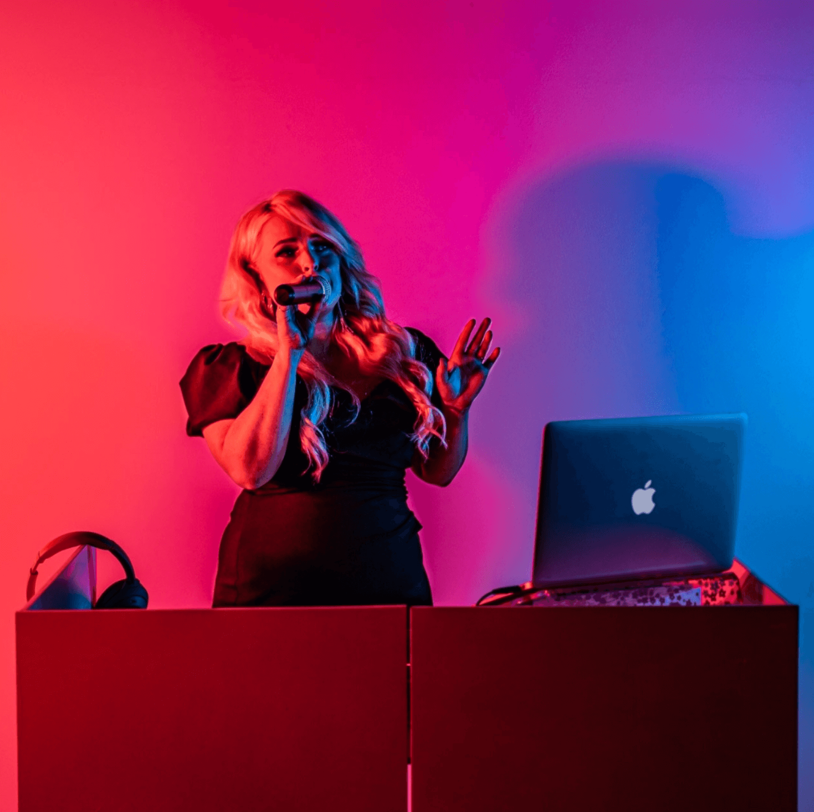 Woman singing into microphone with laptop, headphones, and DJ setup, lit by red and blue lights.