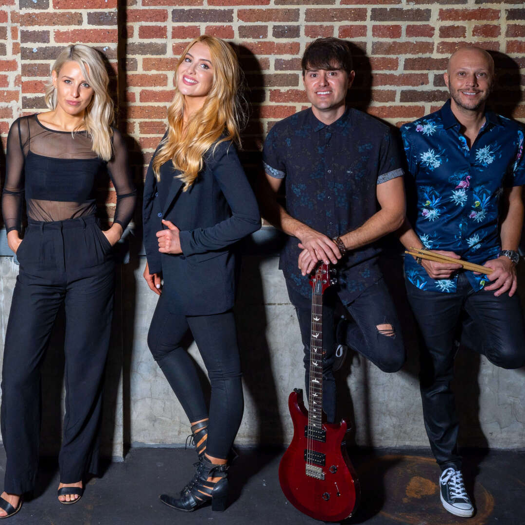 Four band members stand against a brick wall: two women in black outfits, a man holding a red guitar, and one with a shirt.