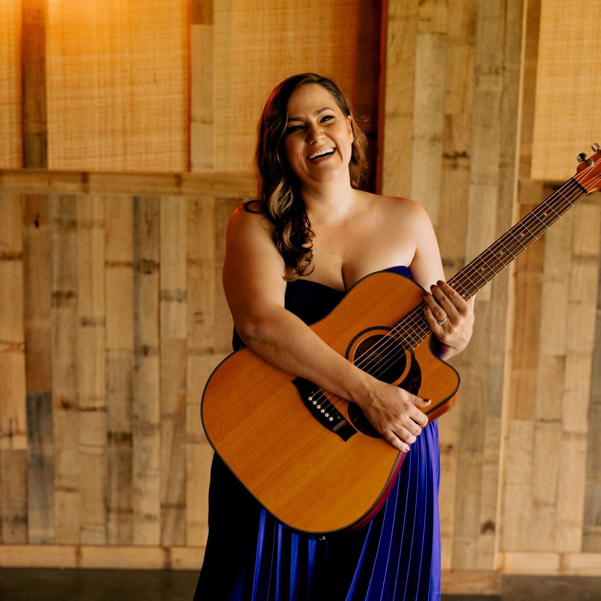 Woman in blue dress plays guitar, smiling. Wooden backdrop.