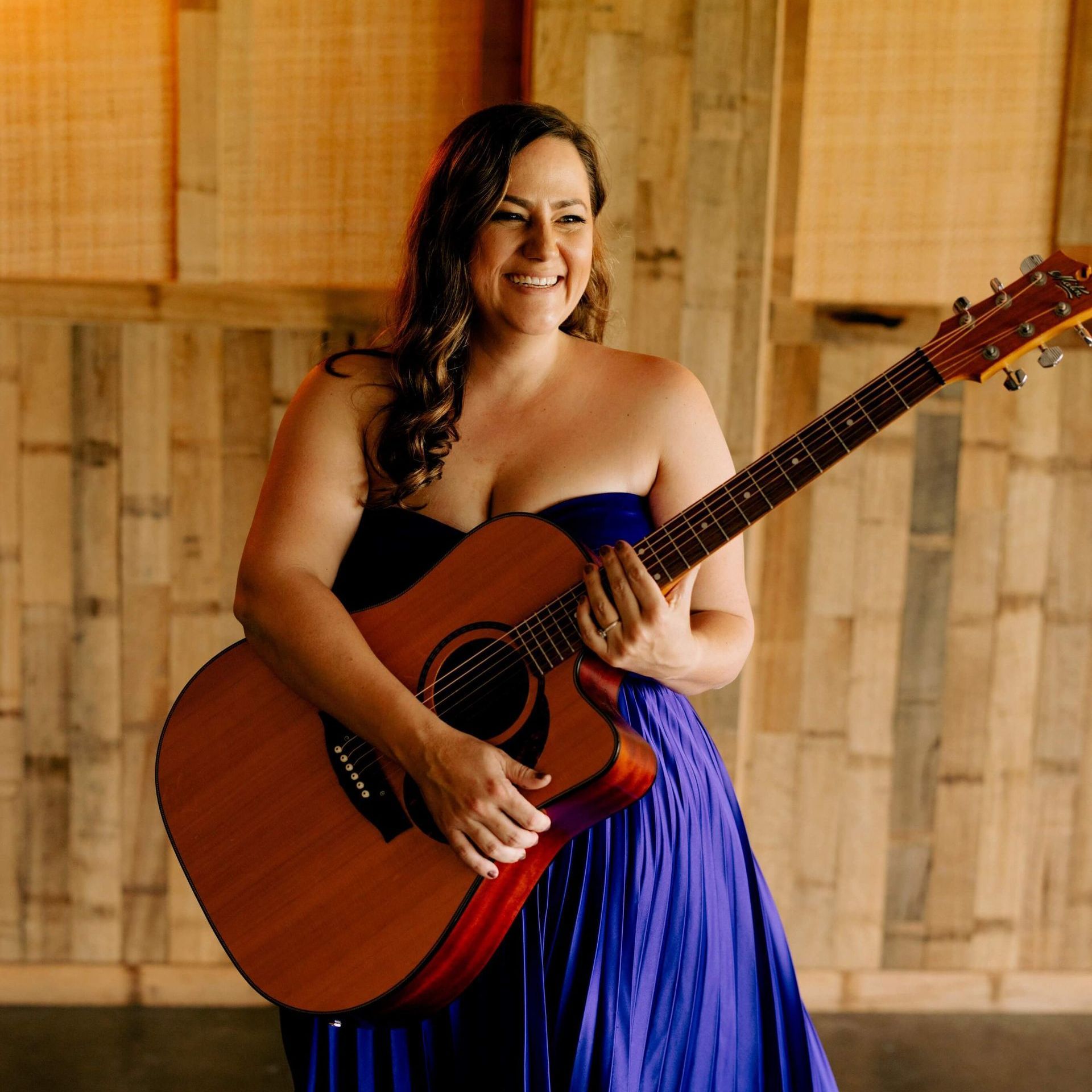 Woman in blue strapless dress, playing acoustic guitar, smiling, wooden background.