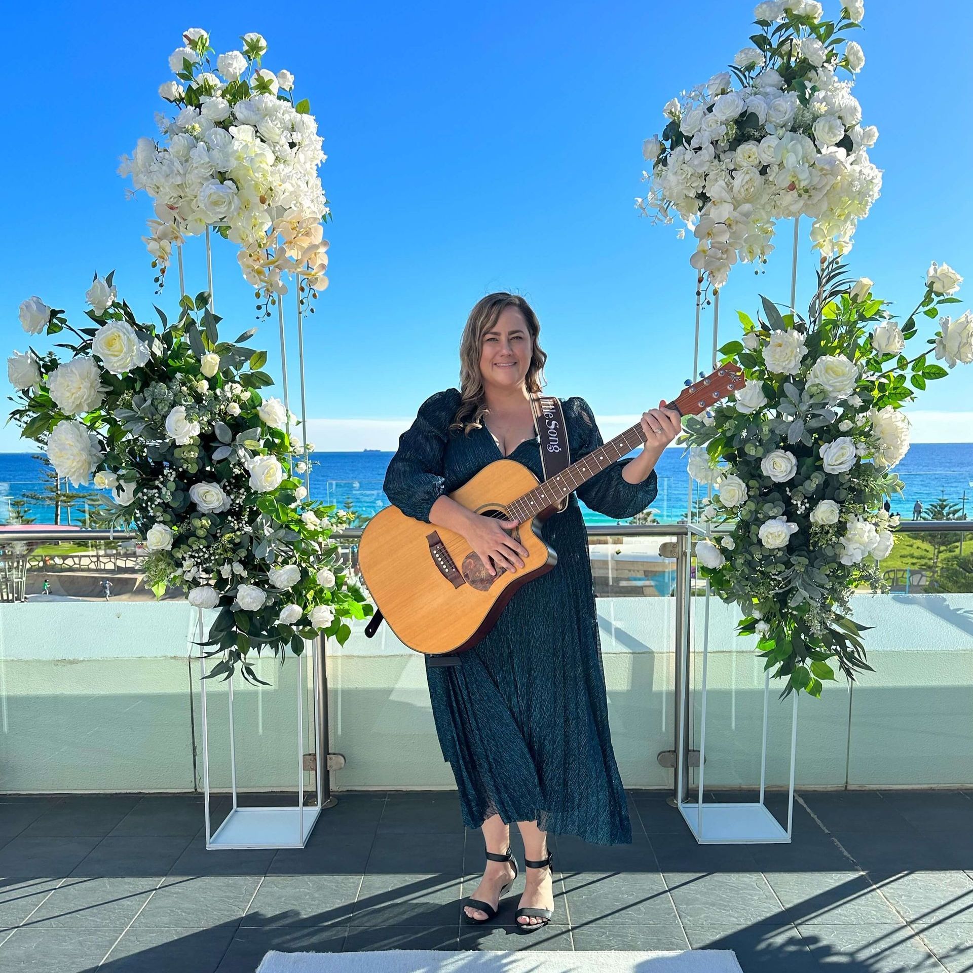 Woman playing guitar outdoors in front of floral arrangements with ocean background.