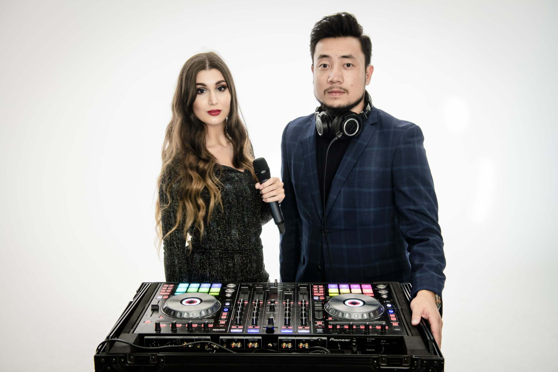 Woman with a microphone and DJ with headphones stand behind a DJ mixer.