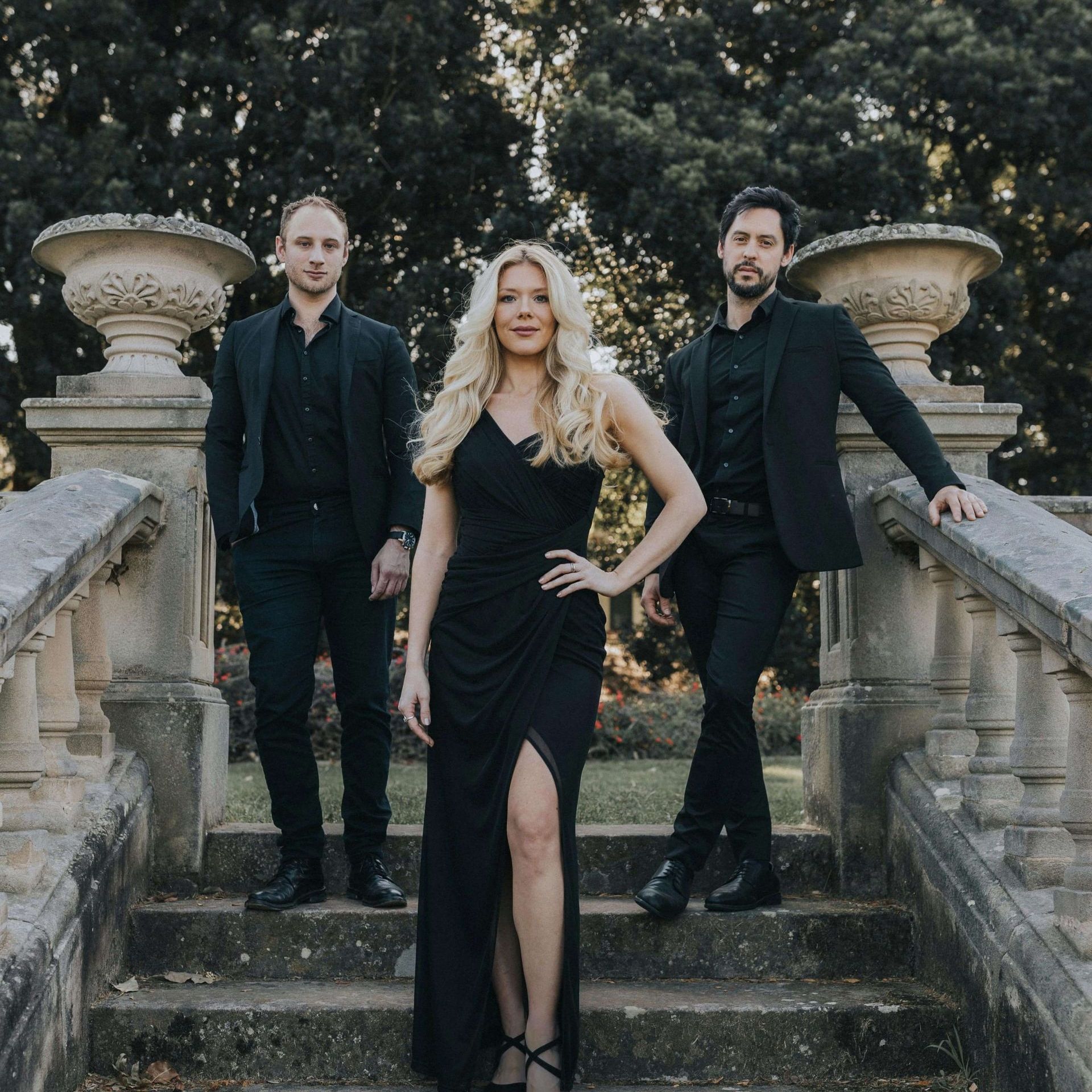 Three people in formal black attire pose on stone stairs flanked by classic urns in an outdoor garden setting.