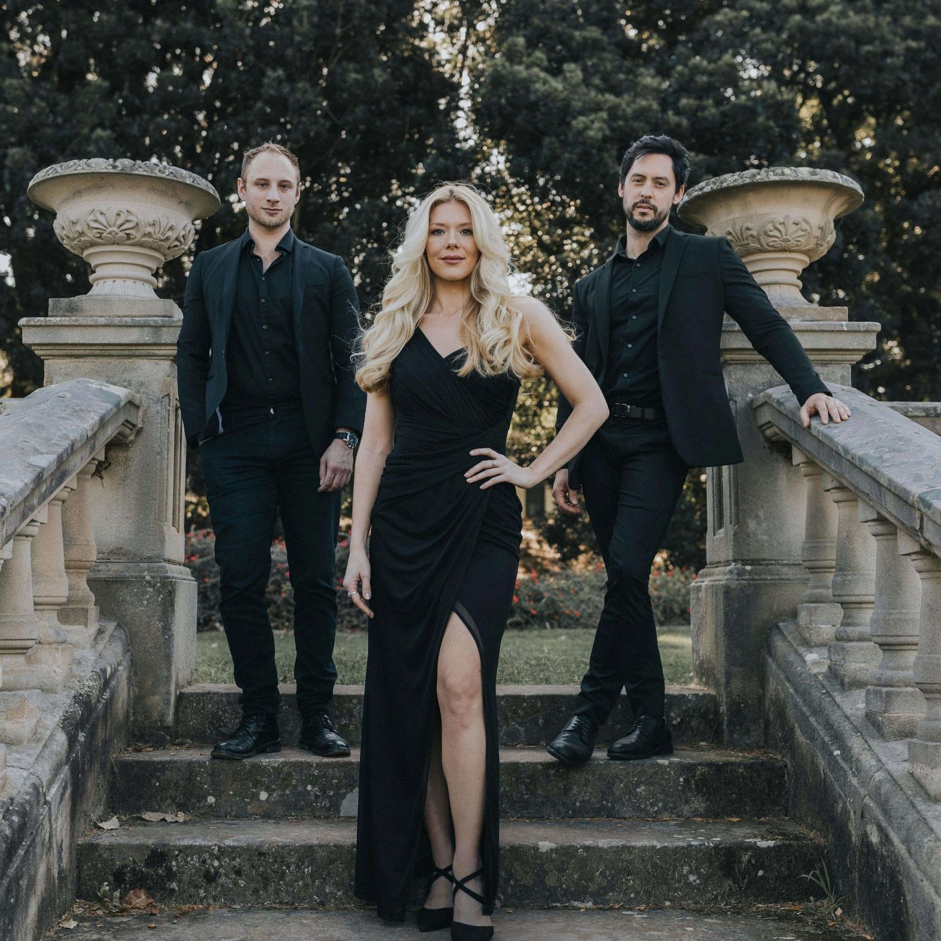 A group of three people dressed in formal black attire stand on a stone staircase surrounded by stone columns and greenery.