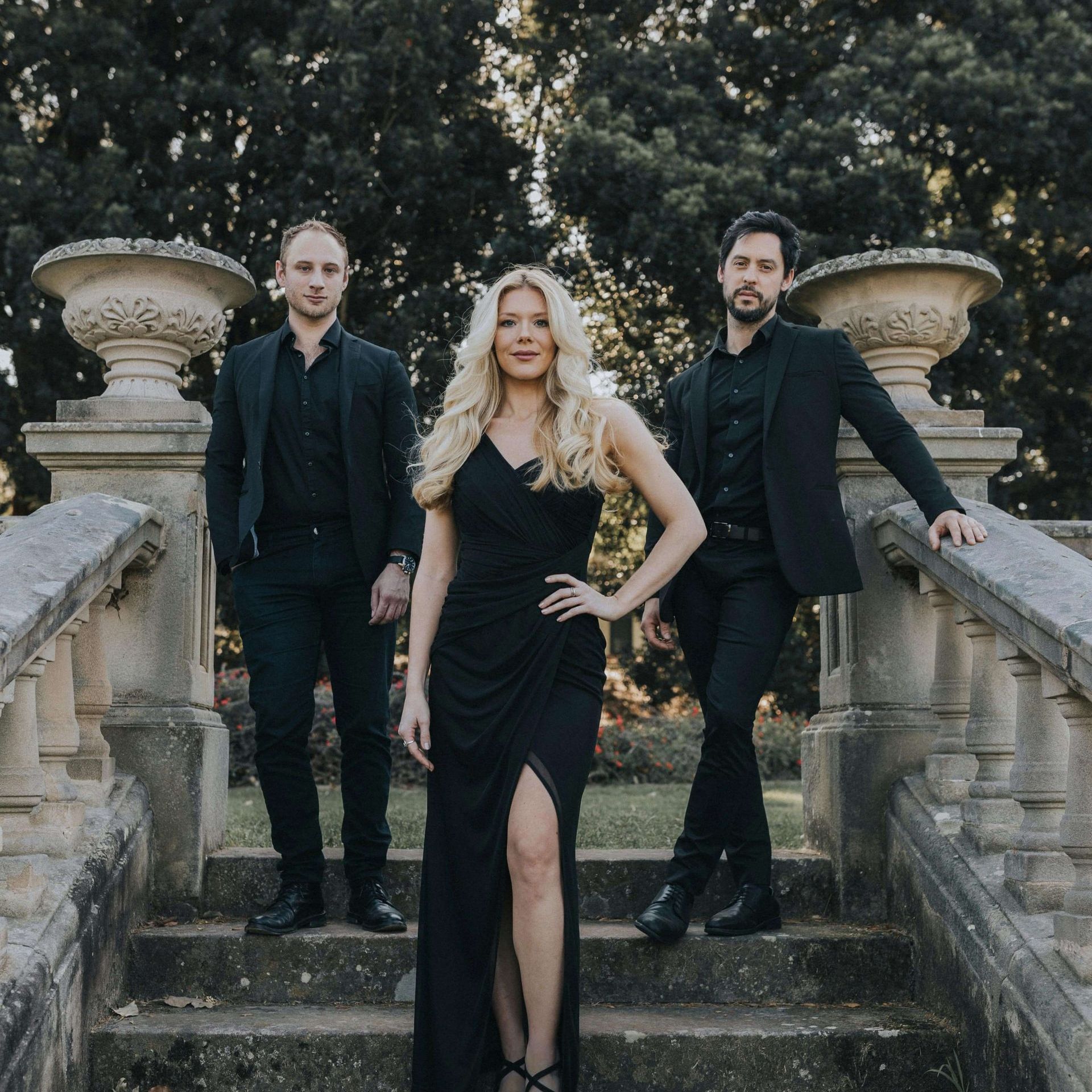 Three people dressed in black formal wear stand on stone stairs outdoors near stone balustrades and urns.