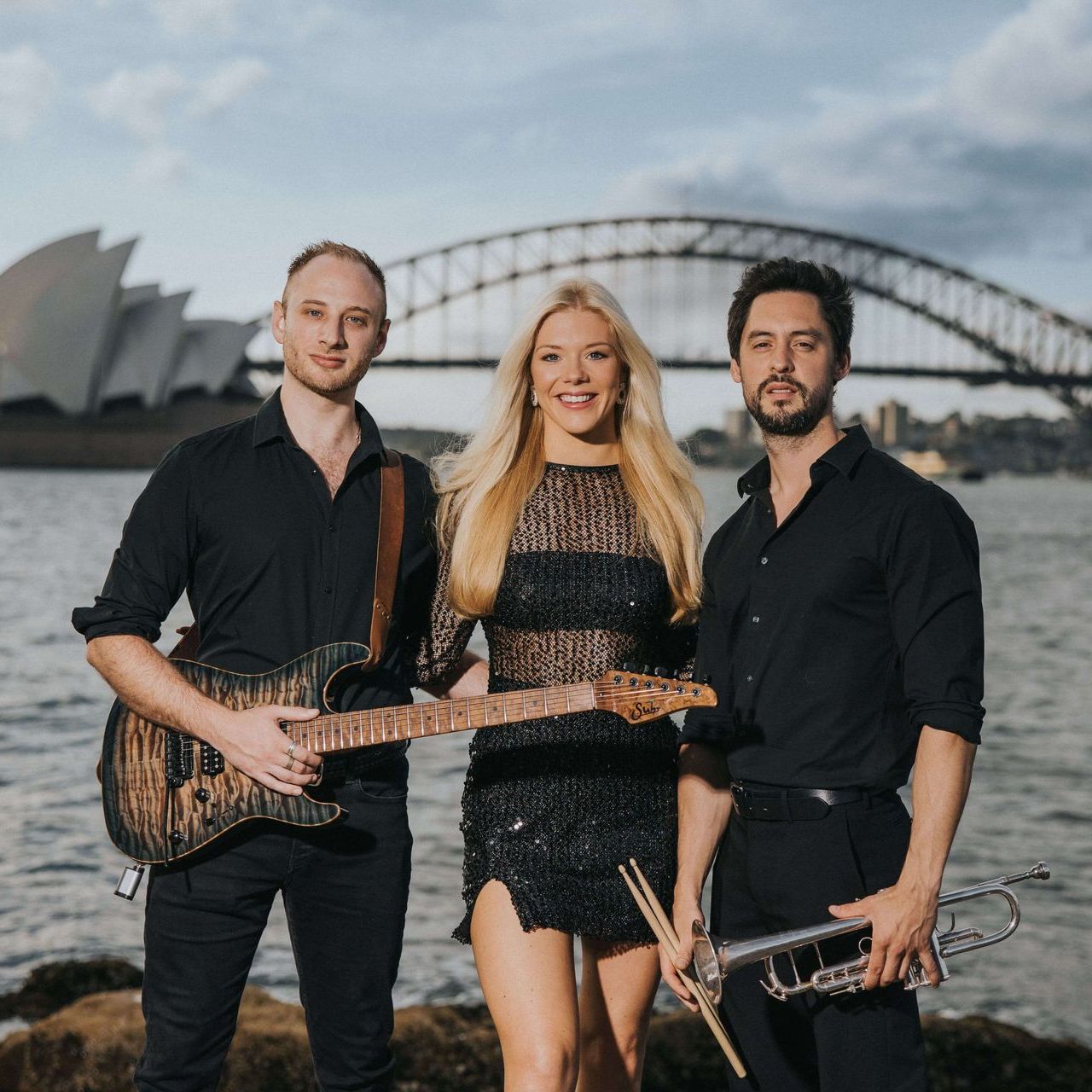 A band of three people holding instruments stand before the Sydney Opera House and Harbour Bridge at dusk.