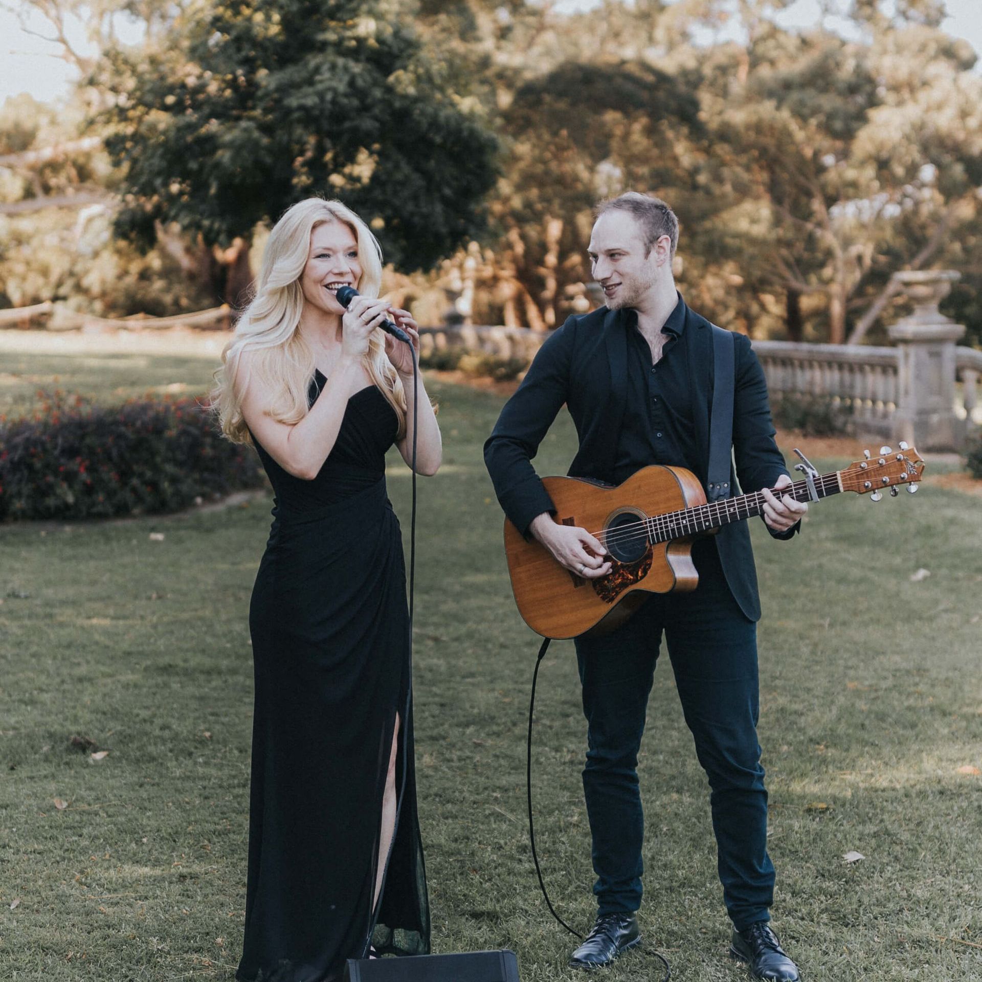 A singer in a black dress and a guitarist in a black outfit perform together on a lawn at an outdoor event.
