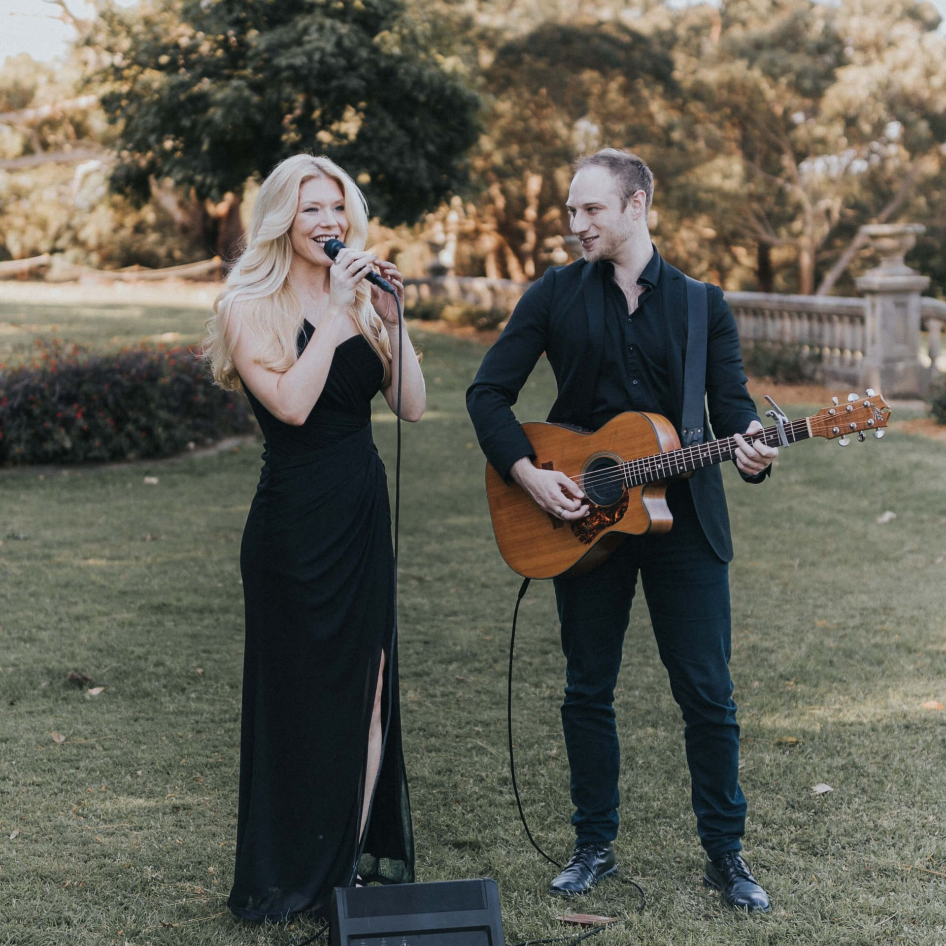 Two musicians perform outdoors, a woman singing beside a man playing acoustic guitar.