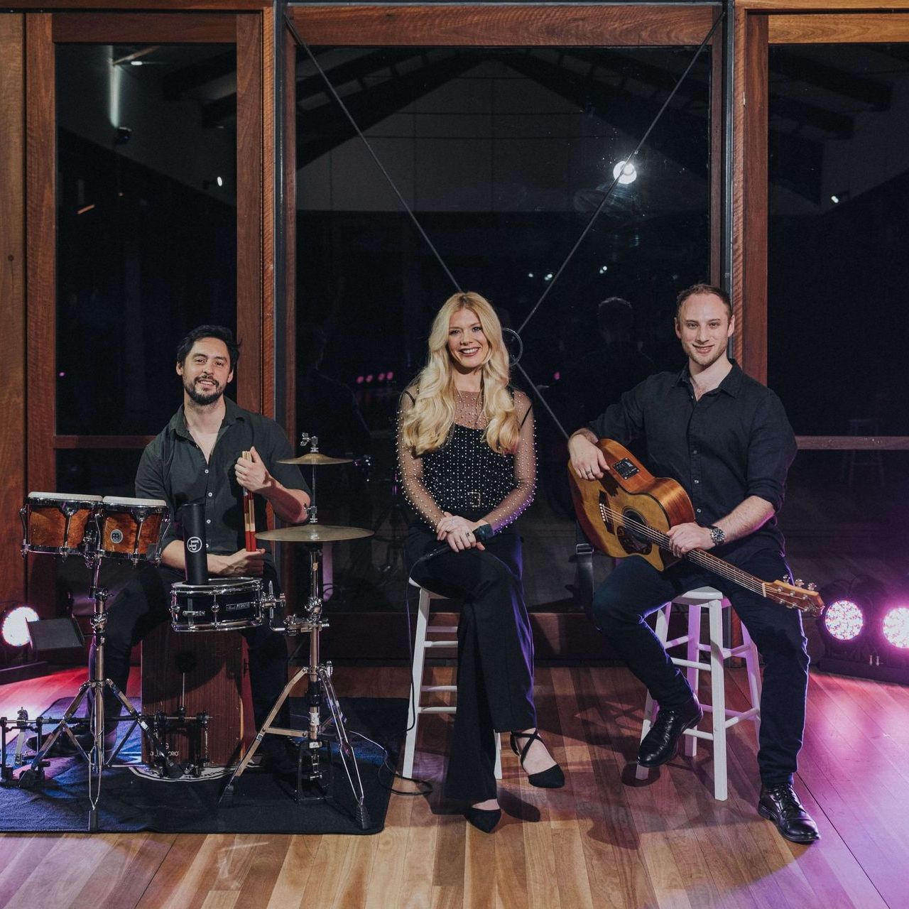 A band of three, including a drummer, a singer in black, and a guitarist, performing on stage in front of window panels.