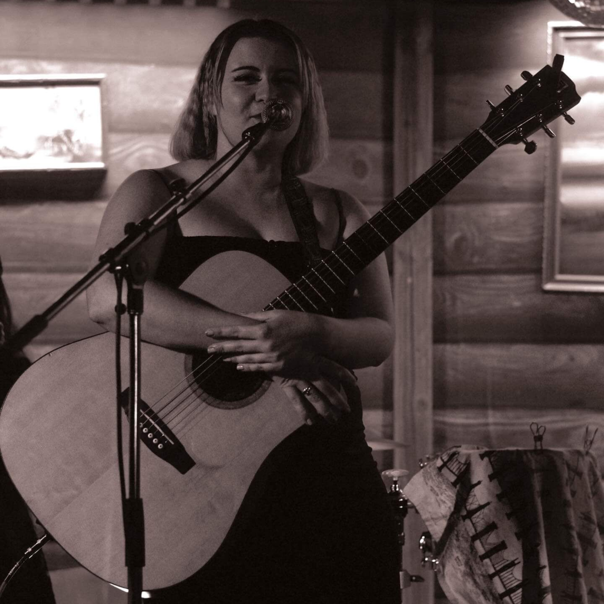 Musician singing into a microphone while holding an acoustic guitar on a wooden stage
