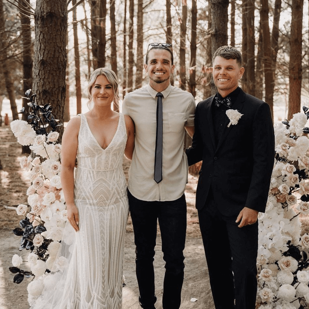 Wedding party poses by floral arch: Bride in lace dress, groom in suit, man in button-down shirt. Outdoor setting.