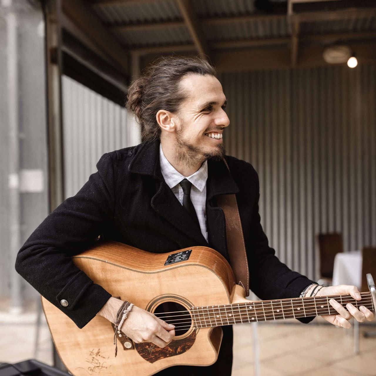 Man in black coat plays acoustic guitar, smiling, outdoors.