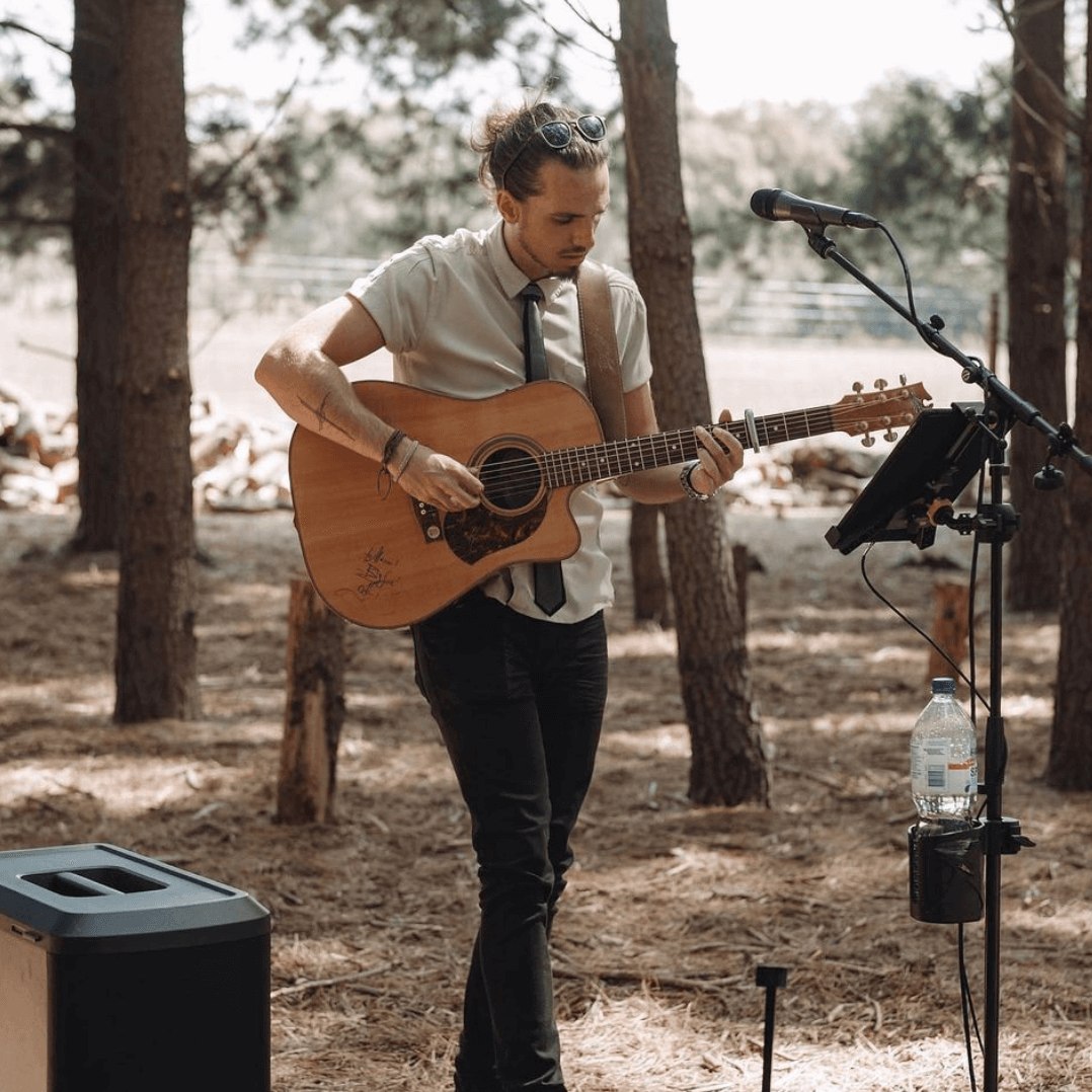 Man playing acoustic guitar outdoors, near microphone and speaker, wearing a tie.