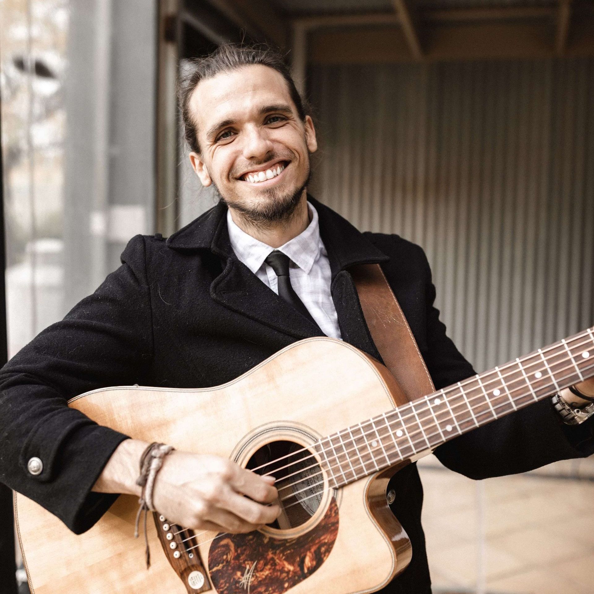 Man with guitar smiling, wearing a coat and tie, playing outdoors.