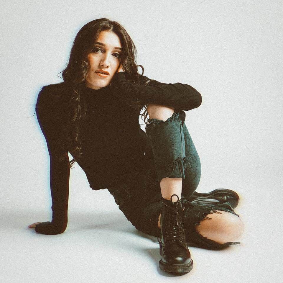 Woman in black turtleneck and ripped jeans, sitting, resting arm on knee, black boots.